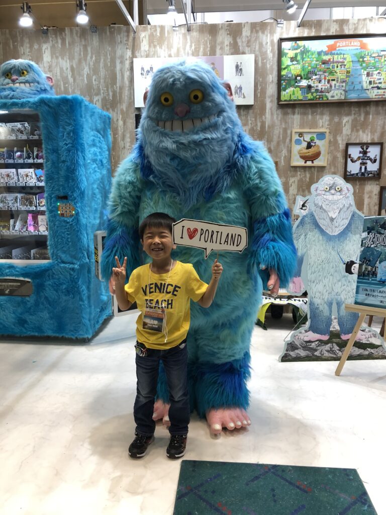 Small child flashing peace sign and holding I Love Portland sign in front of large blue monster mascot